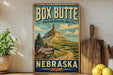 A vintage-style sign for Box Butte State Recreation Area in Nebraska, featuring a mountain landscape and a wooden sign that says "Adventure Awaits".