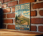 A vintage-style metal sign for the Box Butte State Recreation Area in Nebraska, featuring a mountain landscape and a sign that says "Adventurous Awaits".