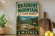 A vintage-style metal sign for Bradbury Mountain State Park in Maine, featuring a scenic landscape with pine trees, mountains, and a lake.