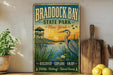 A vintage-style sign for Braddock Bay State Park in New York, featuring an illustration of a bird, a wooden pier, and a sunset over the water.