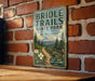 A vintage-style metal sign for Bridle Trails State Park in Washington, featuring a scenic mountain landscape and a trail leading through the woods.