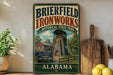 A vintage-style sign for Brierfield Ironworks Historic State Park in Alabama, featuring an illustration of a historic ironworks building and surrounded by a wooden fence.