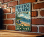 A vintage-style metal sign for Bristol Beach State Park in New York, featuring a beach scene with a sailboat and chairs, mounted on a brick wall.
