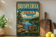 A vintage-style sign for the Brushy Creek State Recreation Area in Iowa, featuring a scenic landscape with a river, mountains, and a pine tree.