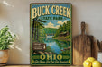 A vintage-style metal sign for Buck Creek State Park in Ohio, featuring a scenic landscape with a lake, forest, and cabin.