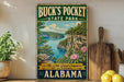 A vintage-style sign for Buck's Pocket State Park, featuring a scenic image of the Little Grand Canyon of Alabama, with a wooden paddle and a potted plant in the foreground.