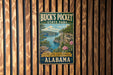 A vintage-style metal sign for Buck's Pocket State Park in Alabama, featuring a scenic landscape with a lake, mountains, and flowers.