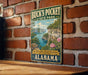 A vintage-style metal sign for Buck's Pocket State Park in Alabama, featuring a scenic landscape with a river and cliffs.