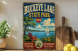 A vintage-style sign for Buckeye Lake State Park, featuring a scenic illustration of a lake, a sailboat, and a wooden dock with a chair.