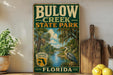 A vintage-style sign for Bulow Creek State Park in Florida, featuring a scenic illustration of a bird and palm trees, with a wooden paddle and a potted plant in the foreground.