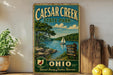 a vintage-style metal sign for Caesar Creek State Park in Ohio, with a scenic illustration of the park's lake and surrounding landscape.