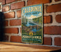 A vintage metal sign for Caledonia State Park in Pennsylvania, featuring a scenic landscape with a river, bridge, and trees.