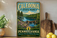A vintage-style metal sign for Caledonia State Park in Pennsylvania, featuring a scenic landscape with a river, bridge, and trees.