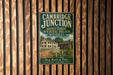 A vintage metal sign for Cambridge Junction Historic State Park in Michigan, featuring an illustration of a log cabin and a dirt road.