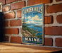 A vintage metal sign for Camden Hills State Park in Maine, featuring a scenic landscape with mountains, ocean, and boats.