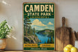 A vintage-style metal sign for Camden State Park in Minnesota, featuring a scenic landscape with pine trees, a lake, and mountains.