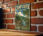 A vintage-style metal sign for Camp Creek State Park in West Virginia, featuring a scenic illustration of a river and forest landscape.