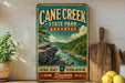 A vintage-style metal sign for Cane Creek State Park in Arkansas, featuring a scenic landscape with a river and mountains.