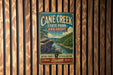 A vintage metal sign for Cane Creek State Park in Arkansas, featuring a scenic landscape with a river and mountains.