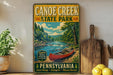 A vintage-style sign for Canoe Creek State Park in Pennsylvania, featuring a scenic image of a canoe on a river surrounded by trees and mountains.