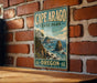 A vintage-style metal sign for Cape Arago State Park in Oregon, featuring a scenic coastal landscape with rocky cliffs, trees, and the ocean.