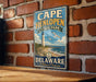 A vintage-style metal sign for Cape Henlopen State Park in Delaware, featuring a lighthouse and beach scene.