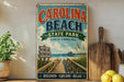 a vintage-style sign for Carolina Beach State Park, with a wooden pier and beach scene, surrounded by various kitchen utensils and a potted plant.