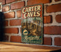 A vintage-style metal sign for Carter Caves State Resort Park in Kentucky, featuring an illustration of a cave and the text "Kentucky Nature's Wonderland Awaits".
