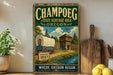 A vintage-style metal sign for Champoeg State Heritage Area in Oregon, featuring an illustration of a covered wagon and a wooden building.