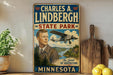 A vintage-style sign for Charles A. Lindbergh State Park in Minnesota, featuring an illustration of a man in a suit and tie standing in front of a scenic landscape with a biplane in the background.