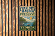 A vintage-style sign for Charlotte Harbor Preserve, Florida, featuring a scenic illustration of the park's natural surroundings.
