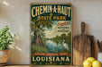 A vintage-style metal sign for Chemin-a-Haut State Park, featuring a lush landscape with a bird and a pond, along with the text "Adventure Nature and History" and "Louisiana".