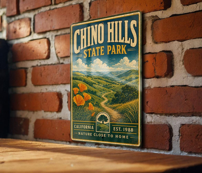 A vintage-style metal sign for Chino Hills State Park, featuring a scenic landscape with a winding path and orange flowers, mounted on a brick wall.