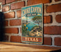 A vintage-style metal sign for Choke Canyon State Park in Texas, featuring a scenic landscape with a lake, mountains, and cacti.