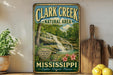 A vintage-style sign for Clark Creek Natural Area in Mississippi, featuring a waterfall and lush greenery.
