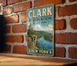 A vintage metal sign for Clark Reservation State Park in New York, featuring a scenic landscape with a lake and mountains.