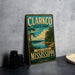 A vintage metal sign for Clarkco State Park, featuring a scenic landscape with a lake and forest, is displayed on a gray countertop next to some black camera lenses.