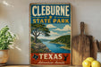 A vintage-style sign for Cleburne State Park, featuring a scenic landscape with a river, trees, and mountains.