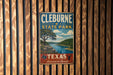 A vintage metal sign for Cleburne State Park, featuring a scenic landscape with a river and trees, mounted on a wooden wall.