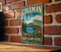 A vintage metal sign for Coleman State Park in New Hampshire, featuring a scenic landscape with pine trees, a lake, and mountains.