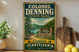 A vintage-style sign for Colonel Denning State Park, featuring a scenic illustration of a forest and a lake, is displayed on a wooden shelf next to a potted plant and a wooden cutting board.