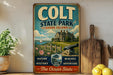 A vintage-style metal sign for "Colt State Park, Rhode Island" featuring a house, a sailboat, and a lighthouse, with the text "NATURE, HISTORY, BEACHES, ADVENTURE - The Ocean State" and "CORTON, RI" on it. The sign is displayed on a shelf next to a potted plant and a wooden cutting board, with a lemon and a knife visible in the background.