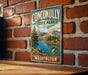 A vintage metal sign for Conconally State Park in Washington, featuring a scenic landscape with mountains, a lake, and pine trees.