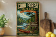 A vintage-style metal sign for Cook Forest State Park in Pennsylvania, featuring a scenic image of a river and pine trees.