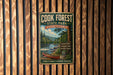 A vintage-style metal sign for Cook Forest State Park in Pennsylvania, featuring a scenic river and pine trees, is displayed on a wooden wall.