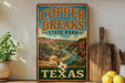 A vintage-style sign for Copper Breaks State Park in Texas, featuring a scenic landscape with a river and mountains.