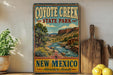 A vintage-style metal sign for Coyote Creek State Park in New Mexico, featuring a scenic landscape with a river and mountains.