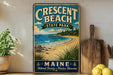 A vintage-style metal sign for Crescent Beach State Park in Maine, featuring a beach scene with a rocky coastline, pine trees, and a blue sky with clouds.