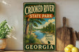 A vintage-style sign for Crooked River State Park in Georgia, featuring a scenic illustration of a river surrounded by lush greenery.