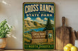 A vintage-style sign for Cross Ranch State Park in North Dakota, featuring a scenic landscape with mountains, a lake, and a wooden signpost.